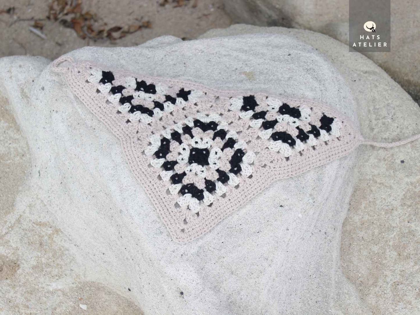 Crocheted triangle bandana with black and white pattern on a stone surface, brand 'Hats Atelier' visible.
