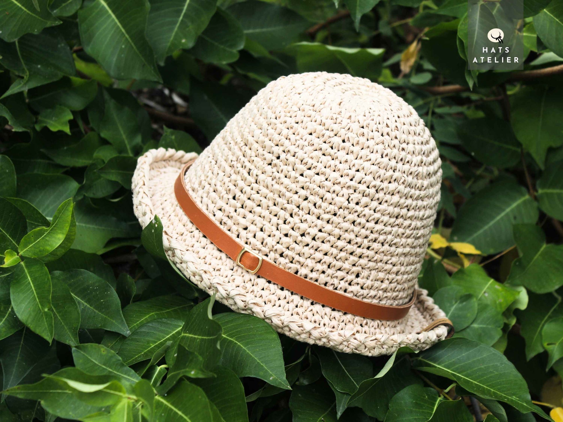 Natural crochet raffia hat with medium brim and leather band on green foliage background